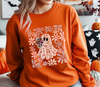 Person wearing an orange sweatshirt with a ghost and floral design, sitting in a room decorated for Halloween.